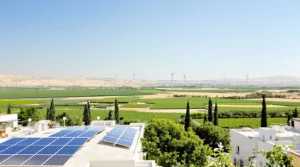 נוף אנרגיה מתחדשת בישראל: גגות עם פאנלים סולאריים בחזית, שדות ירוקים, וטורבינות רוח לבנות על הגבעות ברקע